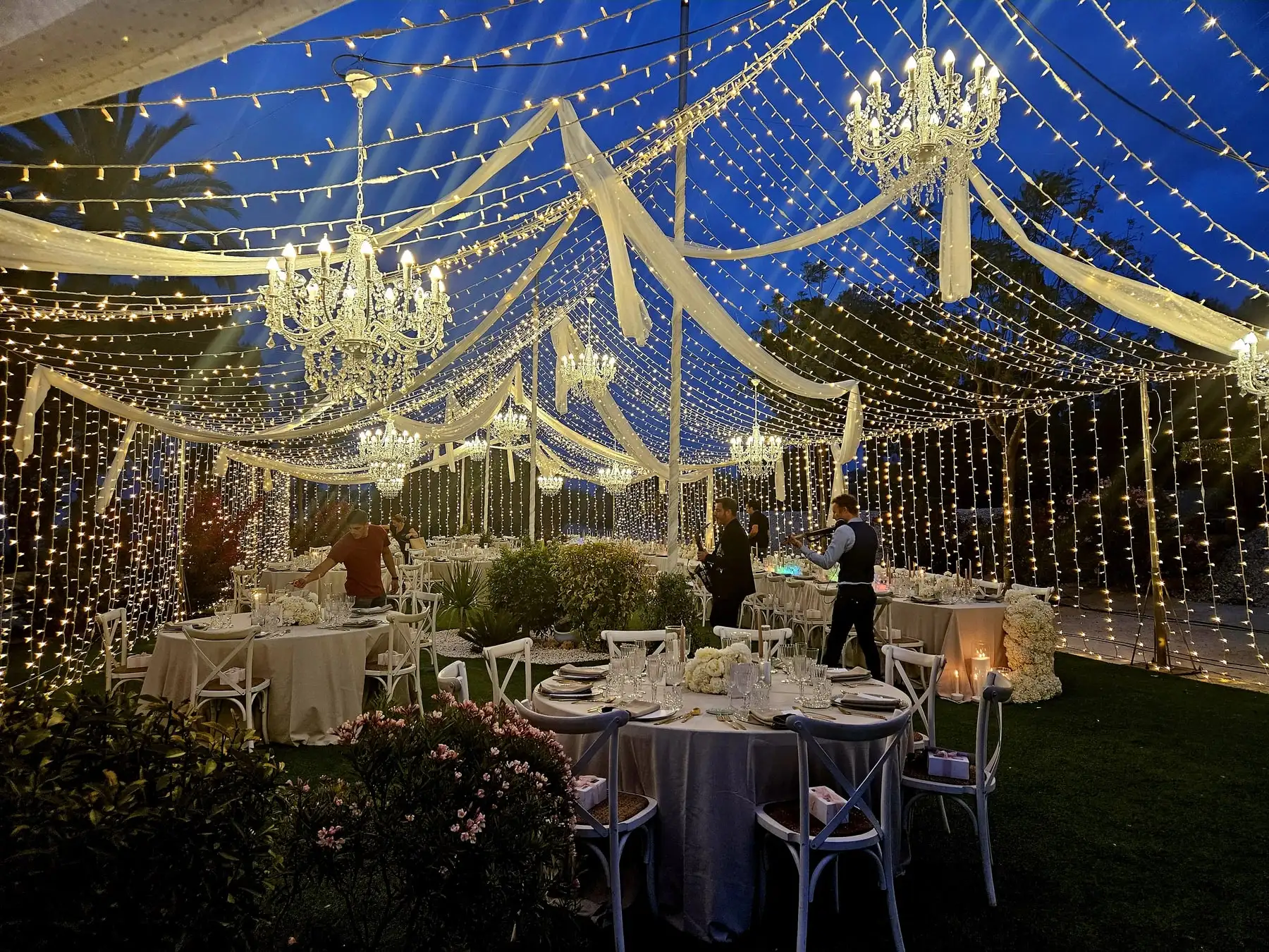 El interior de la carpa por la noche, con lámparas de araña, telas blancas y cortinas de luces sobre las mesas del banquete
