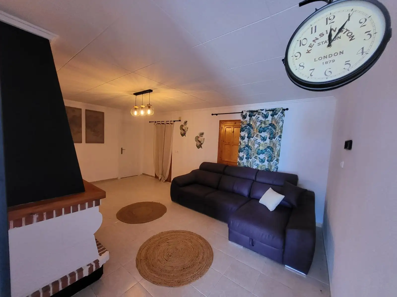 El salón de la Casa Rosa, con chimenea, un sofá oscuro, alfombras de esparto y un gran reloj de estación