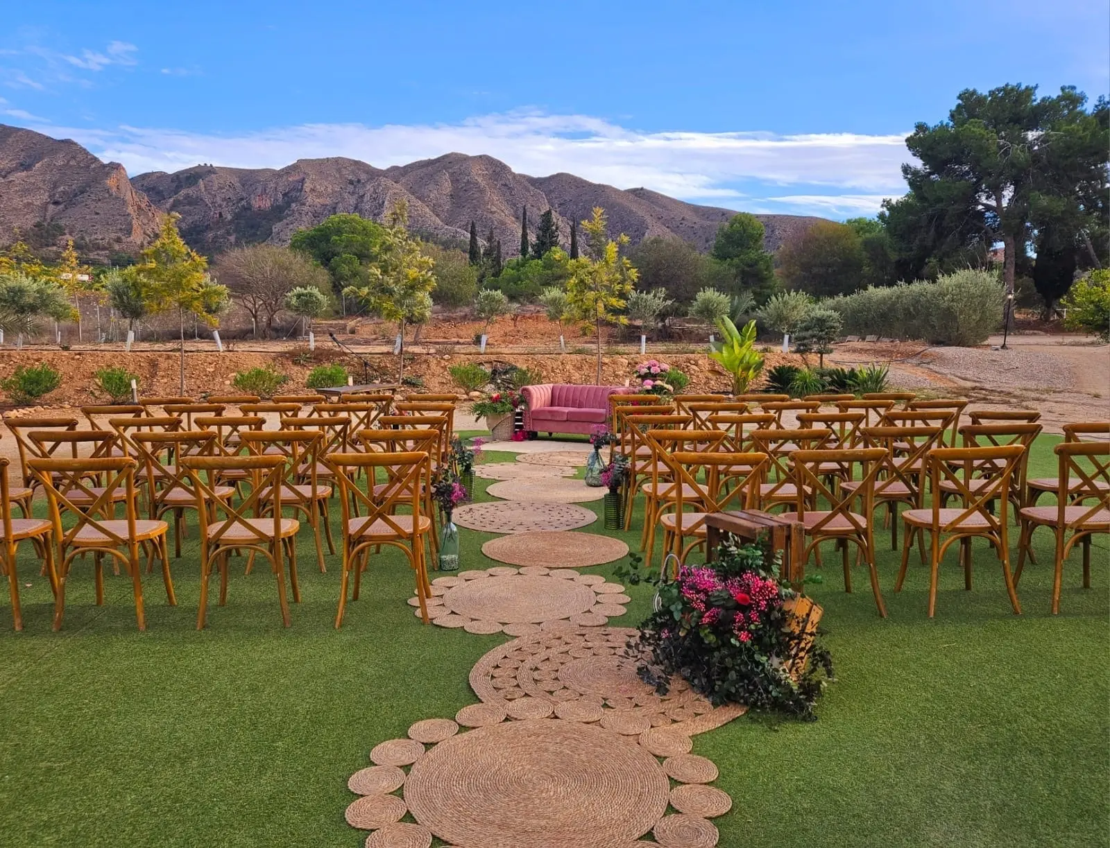 Filas de sillas de madera a ambos lados de un camino de esparto, con un sofá rosa al fondo y la sierra detrás, preparadas para una ceremonia al aire libre