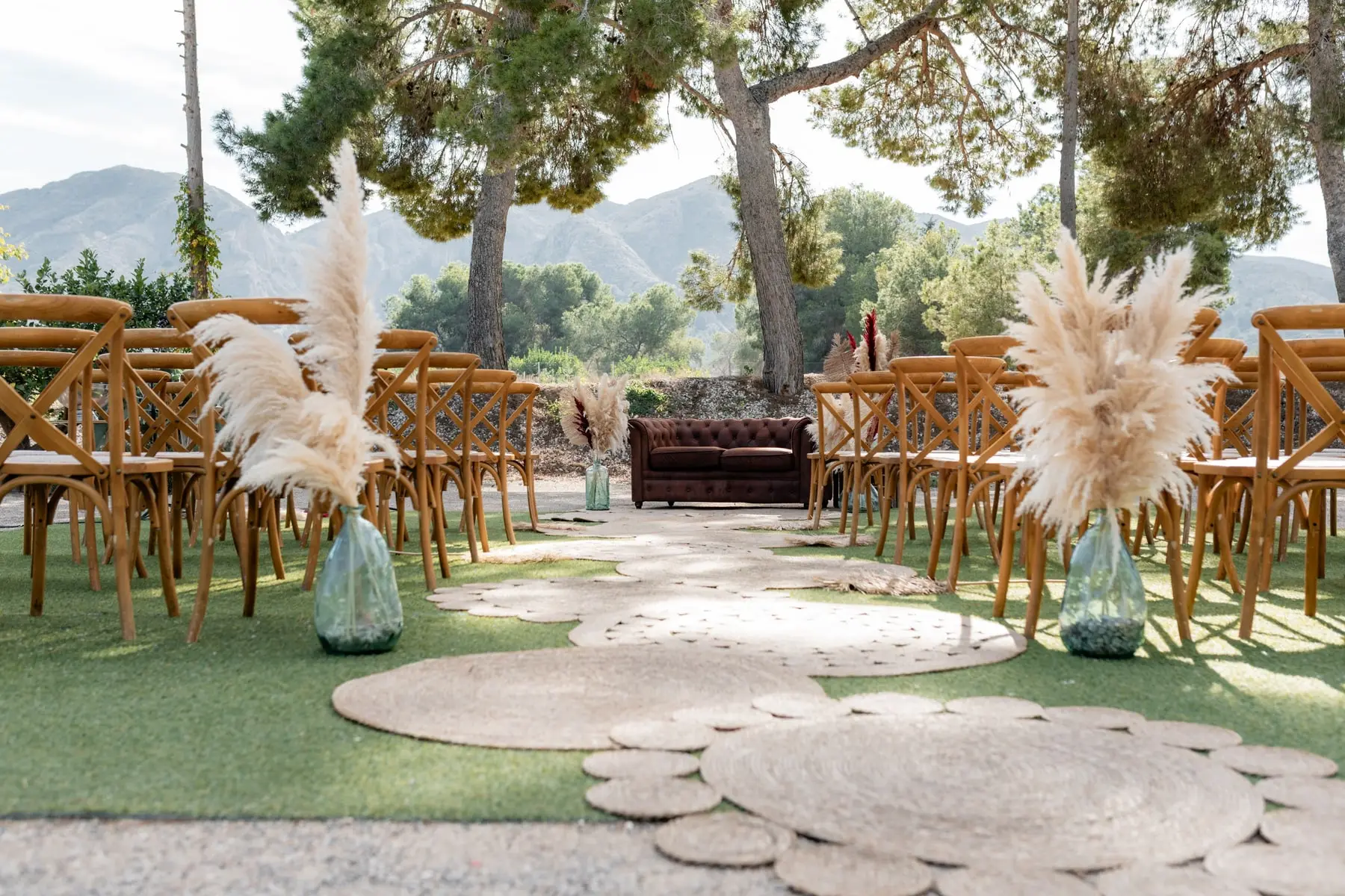 Sillas de madera con plumeros de hierba de la pampa entre pinos, un sofá de cuero al fondo y la sierra detrás, listas para una ceremonia
