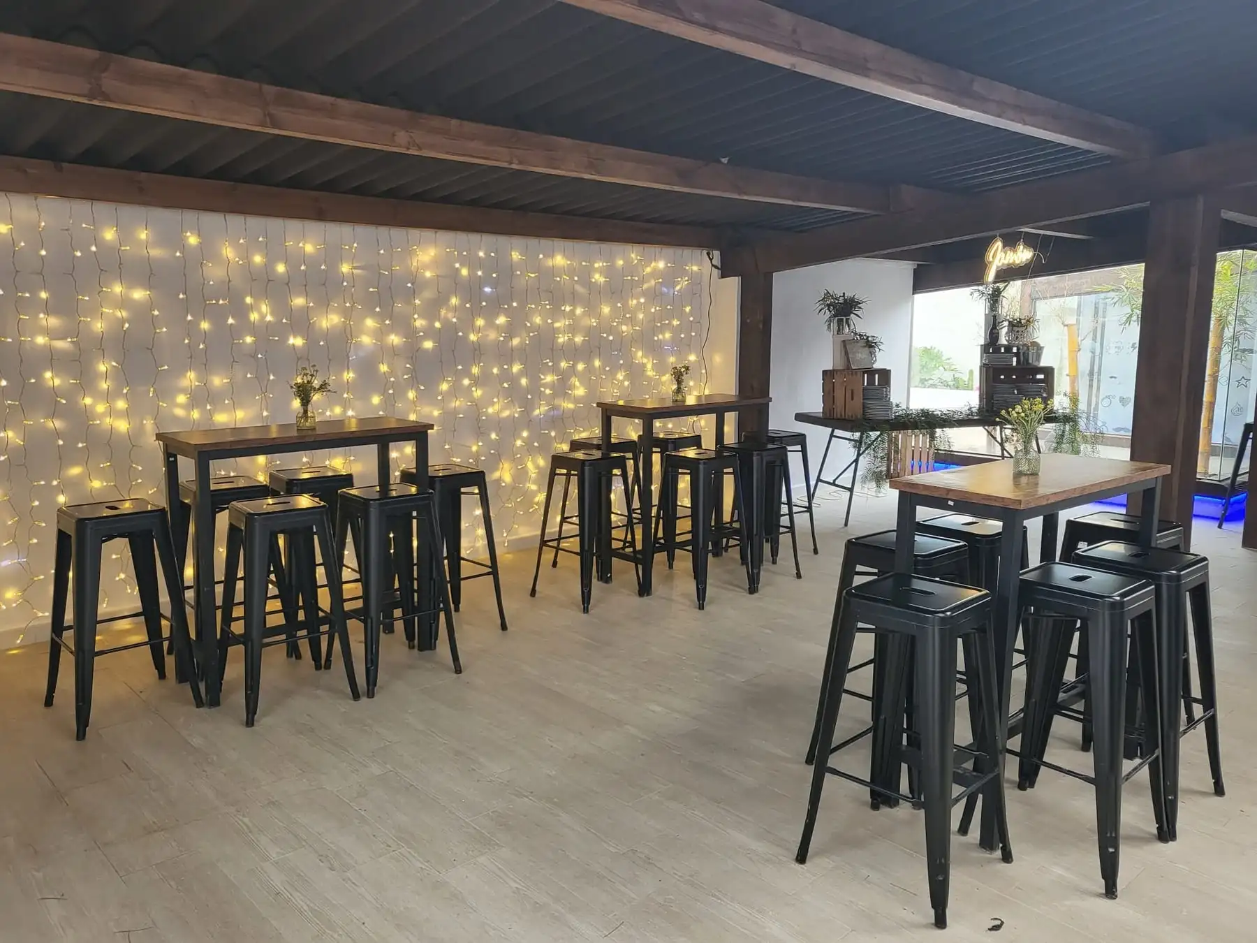 Mesas altas con taburetes negros ante una pared de luces cálidas, en el cobertizo