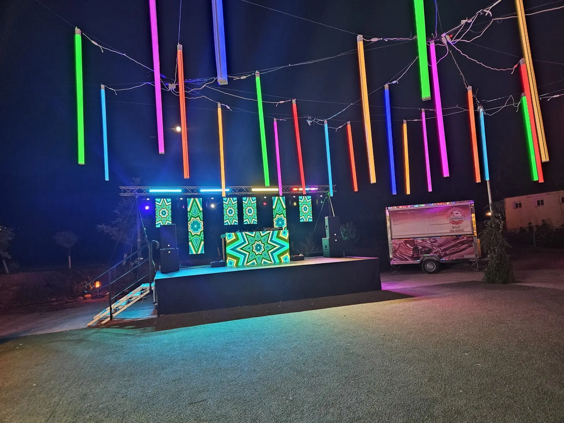 El escenario del DJ de noche, con tubos de luz de colores colgados sobre él y la pista al aire libre en primer plano